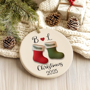 May include: A round Christmas ornament with a gold string. The ornament features a red and green stocking design with the text "Christmas 2025". The ornament also has the letters "B" and "L" with a heart symbol.