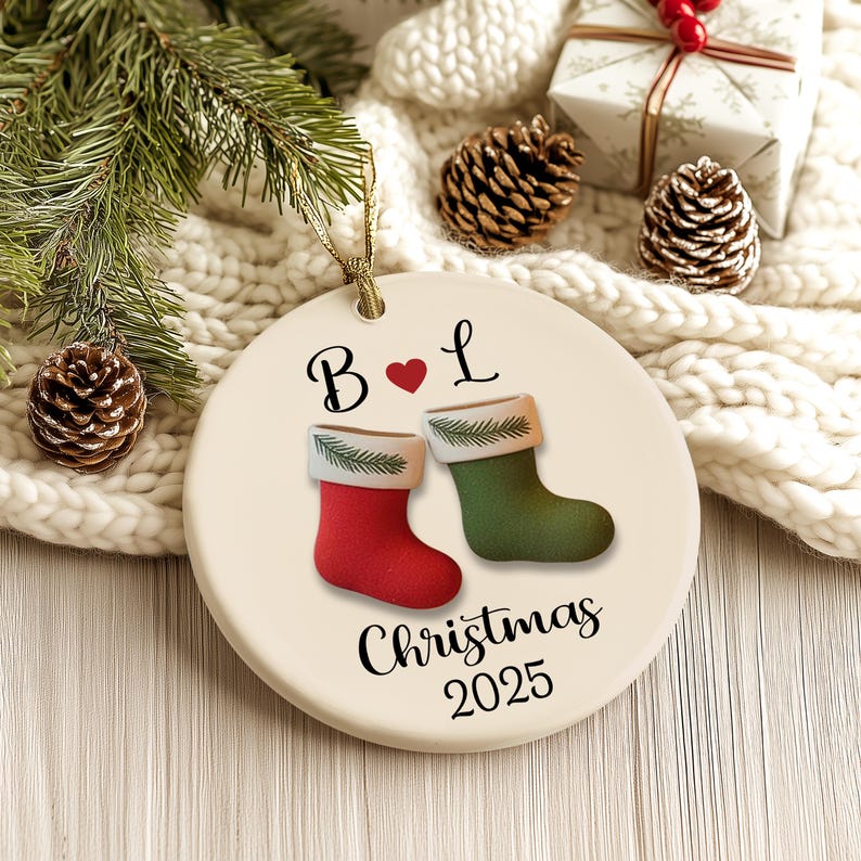 May include: A round Christmas ornament with a gold string. The ornament features a red and green stocking design with the text "Christmas 2025". The ornament also has the letters "B" and "L" with a heart symbol.