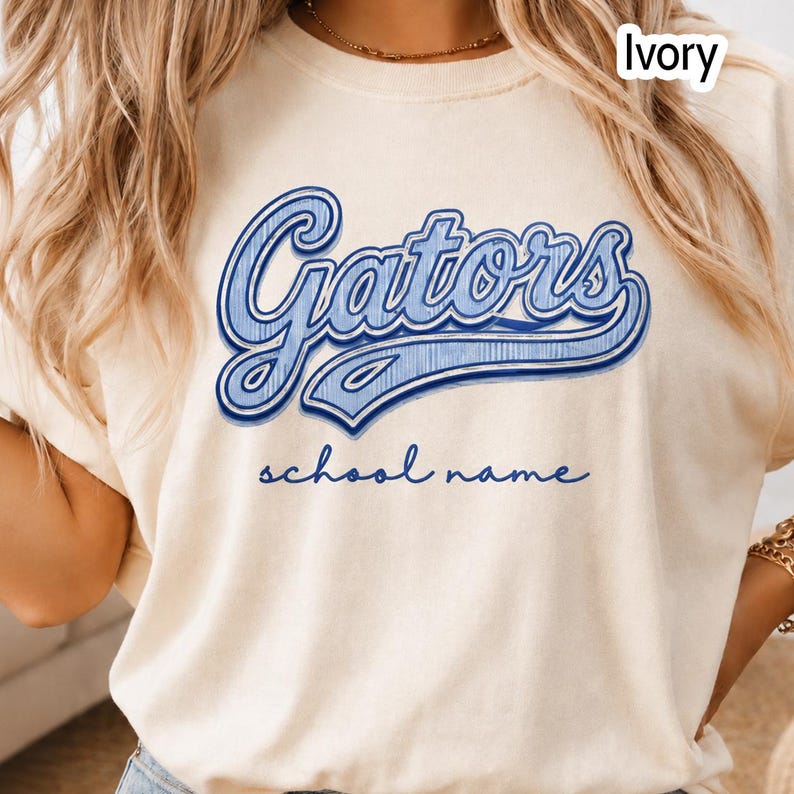 May include: An ivory t-shirt featuring the word Gators in a large, blue, script-style font with a textured, distressed appearance. Below the main text, the words school name are printed in a smaller, simple blue cursive font.