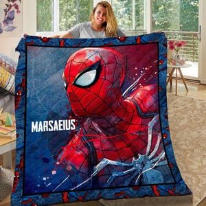 Puede incluir: Un edredón de Spider-Man azul y rojo con un gran gráfico de la cara del superhéroe. El edredón tiene un borde azul con diseños repetidos de Spider-Man y el nombre "MARSAEIUS" en blanco.