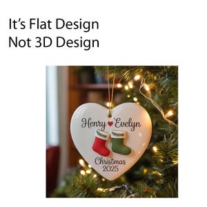 May include: A white heart-shaped Christmas ornament with the names "Henry" and "Evelyn" and the year "2025" printed on it. The ornament features red and green stocking designs. It is hanging on a Christmas tree.