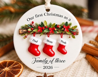 Puede incluir: Un adorno navideño redondo de cerámica blanca con el texto Our first Christmas as a Family of three 2026. Muestra una rama de madera con agujas de pino verdes, bayas rojas y tres calcetines rojos etiquetados como Dad, Mom y Noah.