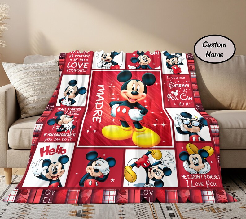 Puede incluir: Una manta roja y blanca con el tema de Mickey Mouse. La manta presenta m&uacute;ltiples im&aacute;genes de Mickey Mouse en varias poses, con la palabra "MADRE" y otro texto. La manta est&aacute; sobre un sof&aacute; beige.