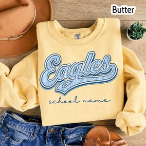 May include: A butter yellow crewneck sweatshirt featuring the word Eagles in a blue and white striped script font with a dark blue outline. Below the word Eagles, the text school name is printed in a dark blue cursive script font.