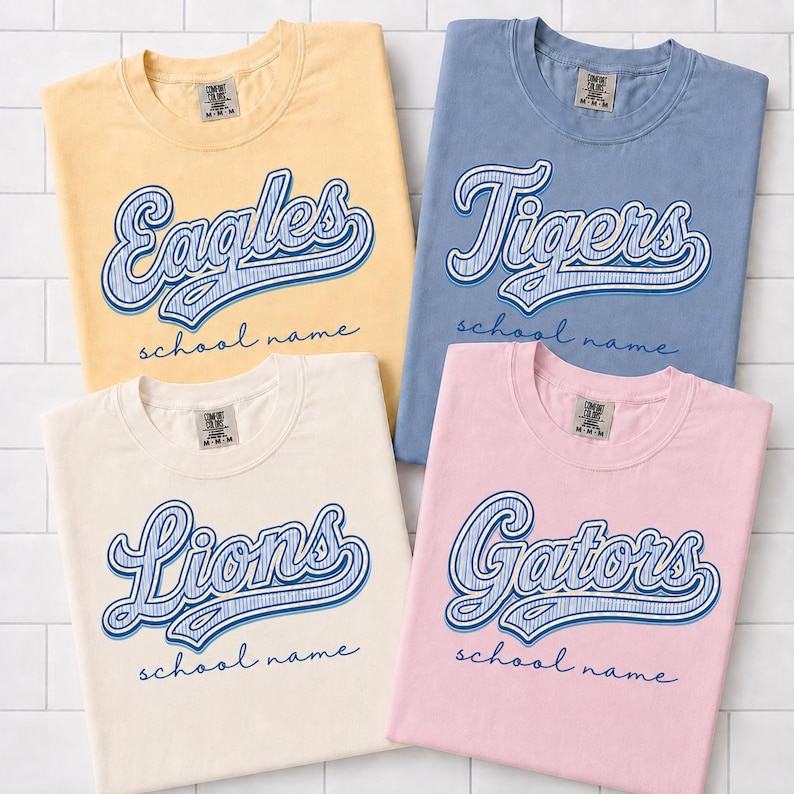 May include: Four short-sleeve crew neck t-shirts in yellow, blue, cream, and pink. Each shirt features a blue script graphic with the words Eagles, Tigers, Lions, or Gators, followed by the text school name in a cursive font below the main graphic.