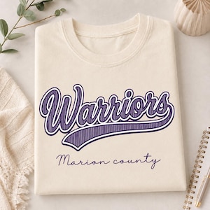 May include: A cream-colored t-shirt featuring the word Warriors in a large, purple, script-style font with a hatched texture. Below the main text, the words Marion county are printed in a smaller, simple, dark purple cursive font.