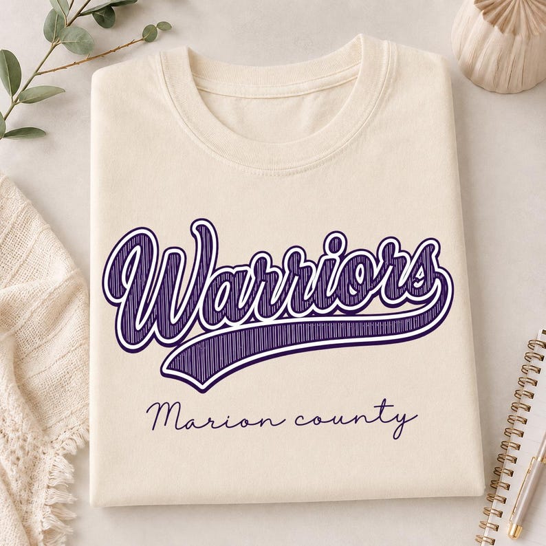 May include: A cream-colored t-shirt featuring the word Warriors in a large, purple, script-style font with a hatched texture. Below the main text, the words Marion county are printed in a smaller, simple, dark purple cursive font.