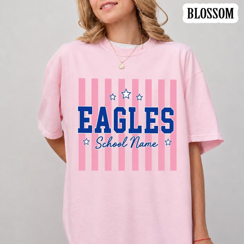 May include: A light pink t-shirt featuring a graphic with vertical pink stripes, the word EAGLES in bold blue block letters, and the text School Name in blue script. The design includes five blue stars and the word BLOSSOM in the top corner.