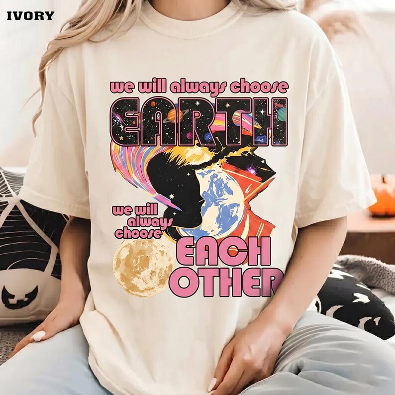 May include: Ivory-colored t-shirt with the text "we will always choose EARTH" and "we will always choose EACH OTHER" in pink and black. The graphic features a silhouette, planets, and a colorful design.