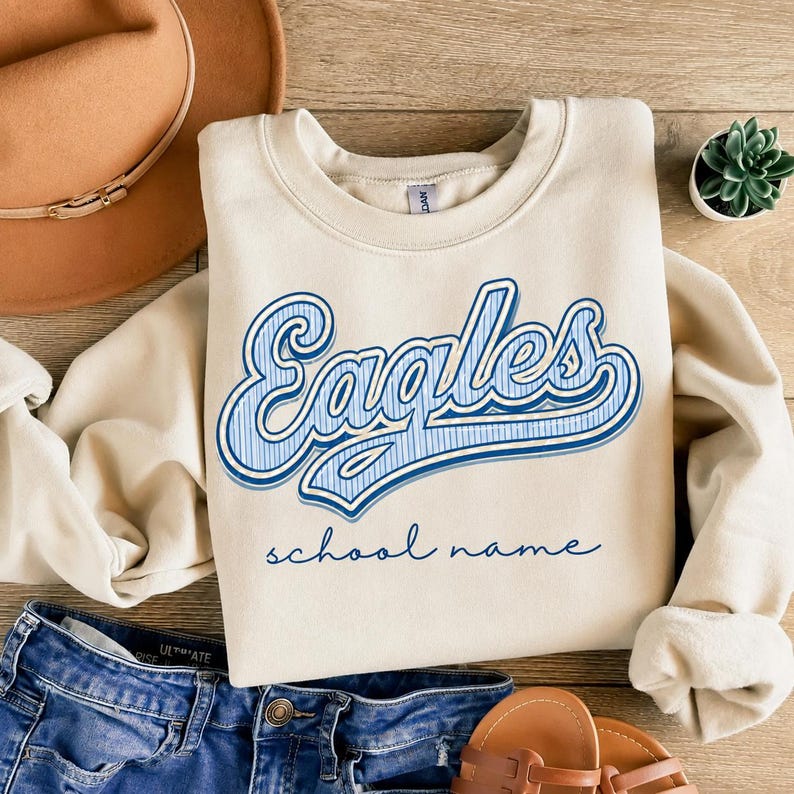 May include: A cream-colored crewneck sweatshirt featuring the word "Eagles" in a blue and white striped font with a blue outline, and the text "school name" in a simple blue script font centered below the main graphic on the chest.