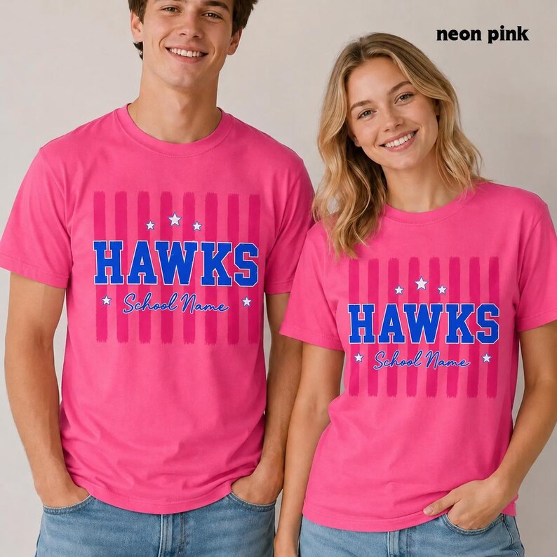 May include: A bright pink short-sleeved t-shirt featuring a vertical striped pattern in a slightly darker shade of pink. The center displays the blue text HAWKS in a bold font, with the blue script text School Name below and white stars.