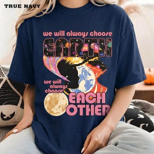 May include: Navy blue t-shirt with the text "we will always choose EARTH, we will always choose EACH OTHER". The graphic features a silhouette, planets, and a colorful rainbow design.