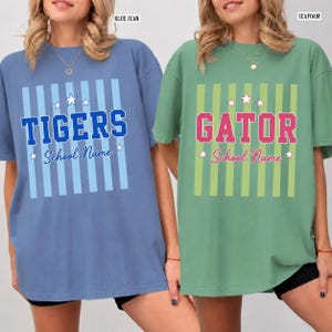 May include: Two short-sleeve crew neck t-shirts. The blue shirt features vertical light blue stripes, white stars, and the text TIGERS School Name. The green shirt features vertical light green stripes, white stars, and the text GATOR School Name.