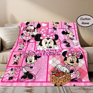 Puede incluir: Una manta rosa de Minnie Mouse con un &aacute;rea de nombre personalizado. La manta presenta m&uacute;ltiples im&aacute;genes de Minnie Mouse en varias poses, sobre un fondo rosa con lunares blancos y un patr&oacute;n de cuadros. La manta est&aacute; sobre un sof&aacute; beige.