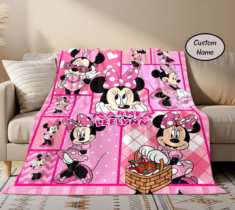 Puede incluir: Una manta rosa de Minnie Mouse con un &aacute;rea de nombre personalizado. La manta presenta m&uacute;ltiples im&aacute;genes de Minnie Mouse en varias poses, sobre un fondo rosa con lunares blancos y un patr&oacute;n de cuadros. La manta est&aacute; sobre un sof&aacute; beige.