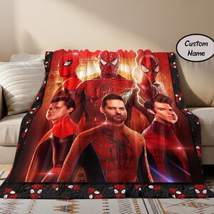 Puede incluir: Una manta roja y negra con tem&aacute;tica de Spider-Man que presenta m&uacute;ltiples personajes de Spider-Man. La manta tiene un borde negro con dise&ntilde;os de m&aacute;scaras de Spider-Man. El fondo es un degradado rojo y naranja.