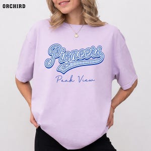May include: A light purple short-sleeve t-shirt featuring the word Pioneers in a large blue script font with white stripes, and the text Peak View in a smaller blue cursive font centered below. The shirt has a crew neckline and a relaxed fit.