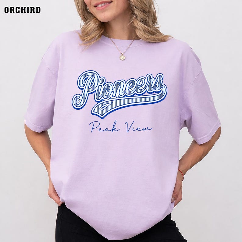 May include: A light purple short-sleeve t-shirt featuring the word Pioneers in a large blue script font with white stripes, and the text Peak View in a smaller blue cursive font centered below. The shirt has a crew neckline and a relaxed fit.