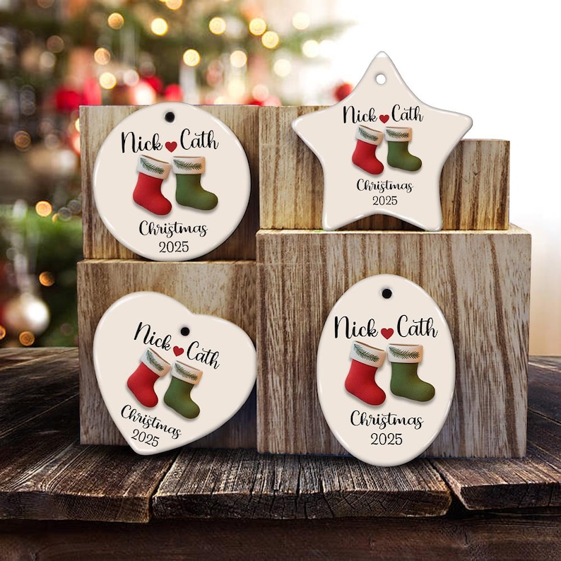 May include: Four Christmas ornaments in various shapes: round, star, heart, and oval. Each ornament features a red and green stocking design, the names "Nick & Cath", and the year "2025". The ornaments are displayed on a wooden surface.