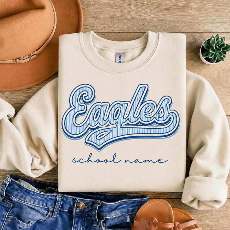 May include: A beige crewneck sweatshirt featuring the word Eagles in a blue and white striped script font, with the text school name printed in a smaller blue cursive font below. The sweatshirt has long sleeves and a ribbed collar.