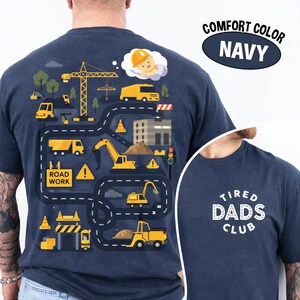 May include: Navy blue t-shirt with a colorful construction-themed graphic on the back. The design includes yellow construction vehicles, a crane, and the words "ROAD WORK." The front of the shirt has the text "TIRED DADS CLUB" in white.