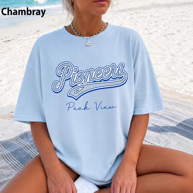 May include: A light blue short-sleeve t-shirt featuring the word Pioneers in a large blue script font with a striped pattern, and the text Peak View in a smaller blue script font underneath. The shirt has a crew neckline and a loose fit.