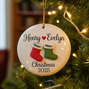 May include: A white ceramic Christmas ornament with the names "Henry" and "Evelyn" above a red heart. Below are two small stockings, one red and one green, and the text "Christmas 2025". The ornament hangs from a gold string.