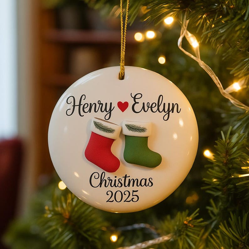 May include: A white ceramic Christmas ornament with the names "Henry" and "Evelyn" above a red heart. Below are two small stockings, one red and one green, and the text "Christmas 2025". The ornament hangs from a gold string.