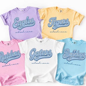 May include: Five short-sleeve crew neck t-shirts in lavender, yellow, pink, white, and light blue. Each shirt features a blue script graphic with the words Eagles, Tigers, Raiders, Gators, or Bulldogs, followed by the text school name.