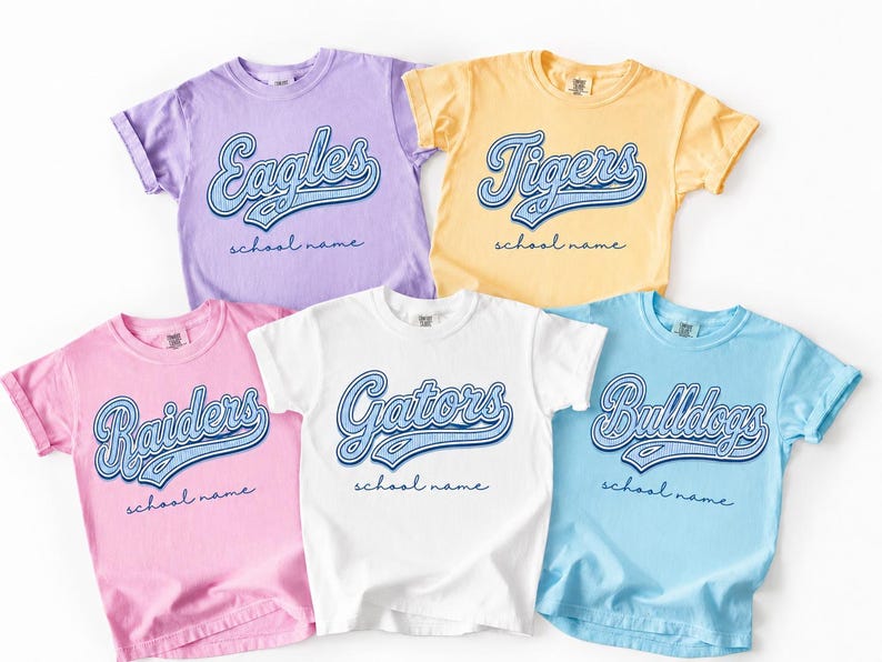 May include: Five short-sleeve crew neck t-shirts in lavender, yellow, pink, white, and light blue. Each shirt features a blue script graphic with the words Eagles, Tigers, Raiders, Gators, or Bulldogs, followed by the text school name.