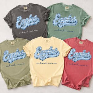 May include: Five short-sleeve crew neck t-shirts in charcoal, sage green, olive green, pale yellow, and faded red. Each shirt features the word Eagles in a blue script font with a white outline, and the text school name in a thin cursive font.