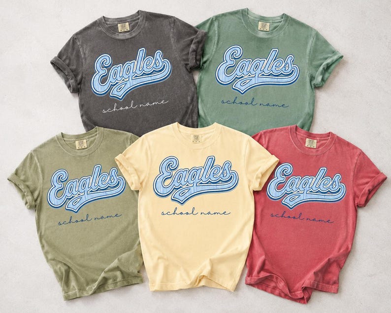 May include: Five short-sleeve crew neck t-shirts in charcoal, sage green, olive green, pale yellow, and faded red. Each shirt features the word Eagles in a blue script font with a white outline, and the text school name in a thin cursive font.