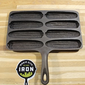 May include: A dark gray cast iron corn stick pan featuring eight individual corn-shaped molds. Beside the pan is a sticker with the text MAGIC CITY IRON EST 2025 and a silhouette of a city skyline. The pan has a single handle with a hole.