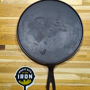 May include: A black cast iron griddle with a single handle and a round cooking surface. The griddle has a smooth cooking surface and a raised edge. A sticker with the text "MAGIC CITY IRON" is next to the griddle.