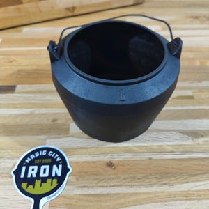 May include: A black cast iron cauldron with a handle, sitting on a wooden surface. A sticker with the text "MAGIC CITY IRON EST 2025" is visible. The cauldron has a wide opening and a tapered base.
