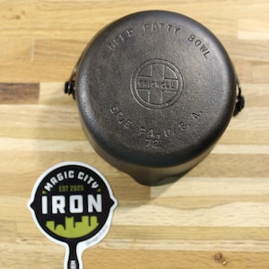 May include: A black cast iron "Beef Patty Bowl" with the text "SHISCLO ERIE PA. U.S.A. 72" embossed on the lid. A "Magic City Iron" sticker is on the wooden surface.