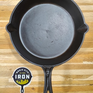 May include: A black cast iron skillet with a long handle and two small spouts. The skillet sits on a wooden surface. A sticker with the text "MAGIC CITY IRON EST 2025" is next to the skillet.