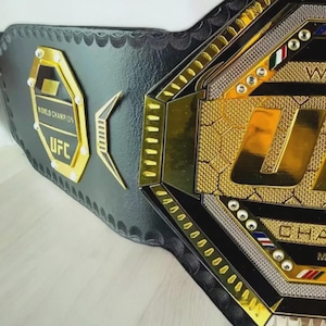以下が含まれることがあります： 黒と金のUFCワールドチャンピオンベルト。ベルトは金のアクセント、UFCのロゴ、そして「ワールドチャンピオン」の文字が特徴です。ベルトは黒革製で、詳細なデザインが施されています。
