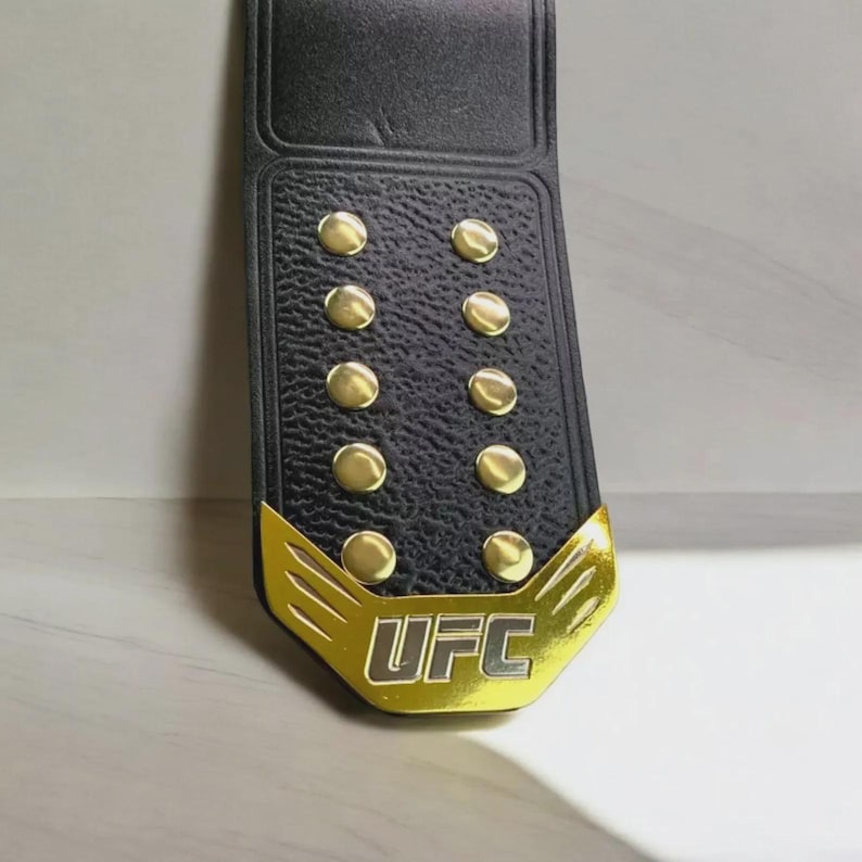 以下が含まれることがあります： 黒い革ベルト、金色のアクセントとスタッド付き。 バックルには金色の「UFC」の文字が入っています。 ベルトは質感のある表面と長方形の形状をしています。
