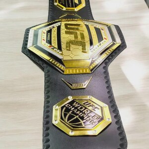 以下が含まれることがあります： 黒革のUFCチャンピオンシップベルト。金と銀のアクセントが施されています。ベルトにはUFCのロゴと「WORLD CHAMPION」と「CHAMPION」の文字が記されています。総合格闘技における勝利の象徴です。