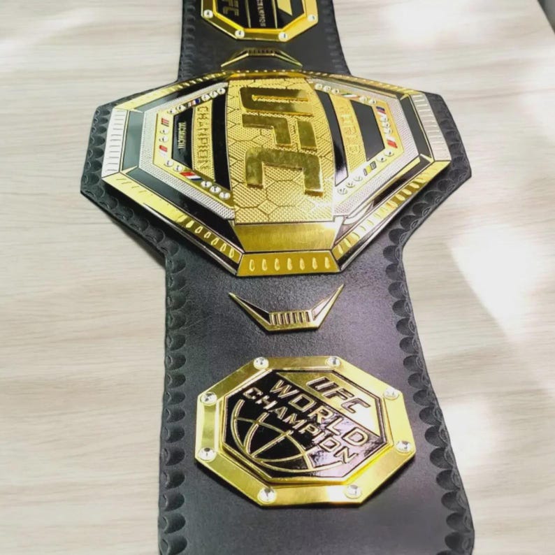 以下が含まれることがあります： 黒革のUFCチャンピオンシップベルト。金と銀のアクセントが施されています。ベルトにはUFCのロゴと「WORLD CHAMPION」と「CHAMPION」の文字が記されています。総合格闘技における勝利の象徴です。