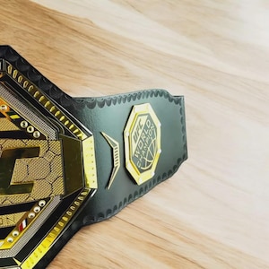 以下が含まれることがあります： UFCワールドチャンピオンベルトのクローズアップ。ベルトは、金と銀のアクセントが付いた黒い革ストラップが特徴です。中央のプレートにはUFCのロゴと「WORLD CHAMPION」の文字があります。