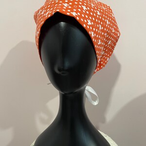 May include: An orange fabric head covering with a white ghost pattern. The head covering is tied in the back with a white ribbon. The head covering is displayed on a black mannequin head.