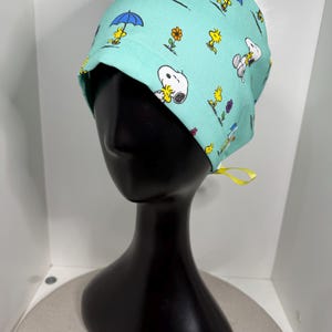 Peut inclure: Une coiffe de chirurgien bleu sarcelle clair avec un motif répété de Snoopy, Woodstock, parapluies et fleurs. Le bonnet est noué dans le dos avec un ruban jaune. Le bonnet est présenté sur une tête de mannequin noire.