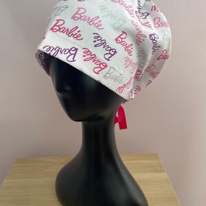 Könnte beinhalten: Eine weiße OP-Haube mit dem Wort "Barbie" in Rosa und Grau bedruckt. Die Haube ist hinten mit einem rosa Band gebunden. Sie wird auf einem schwarzen Schaufensterkopf präsentiert.
