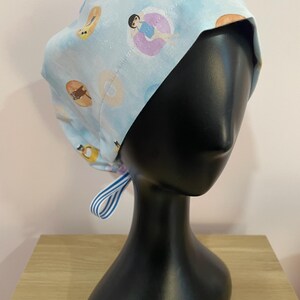 Op de afbeelding: Een lichtblauwe chirurgische scrubcap met een patroon van donuts en mensen in zwembanden. De cap is aan de achterkant vastgebonden met een blauw-wit gestreept lint. De cap wordt getoond op een zwarte mannequinhoofd.