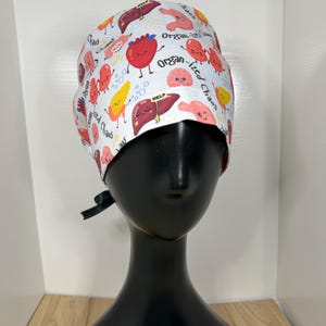 May include: White surgical cap with a repeating pattern of cartoon organs, including hearts, livers, and brains. The cap has the words "Organ-ized Chaos" and "Chaos" printed on it. A black ribbon is tied at the back.
