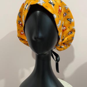 May include: A yellow Halloween-themed scrub cap featuring a repeating pattern of cartoon characters, bats, and spiderwebs. The cap has a black tie in the back and is displayed on a black mannequin head.