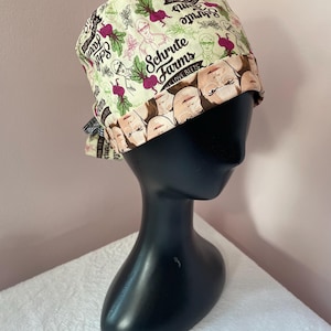 Puede incluir: Un gorro de quirófano beige y verde con un lazo blanco y negro. El gorro presenta un patrón repetido de remolachas, una imagen de dibujos animados de un hombre y el texto "Schrute Farms Love Beets".