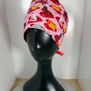 May include: A pink surgical scrub cap featuring a repeating pattern of cats lounging on red hearts. The cats are in various colors, including gray, orange, and brown. The cap has a red tie in the back.